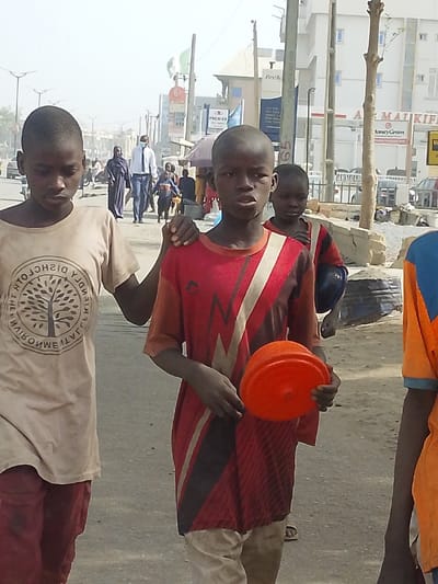 The image depicts an outdoor daytime scene on an urban street in Gandu Albasa, Nigeria. In the immediate foreground, three young males are visible. The leftmost boy, with dark skin and short hair, wears a light brown short-sleeved t-shirt featuring a circular logo with a tree design and mirrored text. He wears dark trousers and looks towards the lower left. The central boy, also with dark skin and short hair, wears a red short-sleeved t-shirt with black, white, and lighter red geometric patterns and light-colored trousers. He holds an orange plastic bowl in his left hand and places his right hand on the left shoulder of the boy to his immediate left. This central boy looks directly towards the viewer. The third boy, partially visible on the right, wears an orange and blue shirt.
In the midground, several pedestrians are present on what appears to be a sidewalk or dirt path adjacent to the street. A man in a blue suit, white shirt, and light blue face mask walks away from the viewer, accompanied by a woman in a dark, flowing garment (abaya/hijab). Other indistinct figures are also walking.
The background reveals a bustling street lined with multi-story buildings. Visible signs include "FirstBank," "PREMIER," "AL MAI KIFIEN," and "MoneyGram." Utility poles, one marked with the number "65," and streetlights are present. Green and white flags, likely Nigerian, are displayed on a pole. Cars are parked or moving in the distance. The atmosphere appears dusty or hazy, suggesting dry weather conditions. Bare tree branches are also visible.