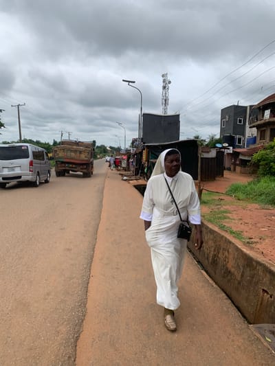 A woman dressed in a white nun's habit with a white veil is walking on the right side of a paved road in Nsugpe, Nigeria. She is wearing dark shoes and carrying a small black purse across her chest. The road is a reddish-brown color and appears to be a main thoroughfare, with a silver van and a large truck carrying dirt traveling in the same direction as the woman. The truck is prominent in the scene, positioned to the left of the woman. On the right side of the road, there are several buildings with corrugated metal roofs, some appearing to be shops or market stalls. The environment suggests a semi-urban setting, with some vegetation visible in the background. The sky is overcast with heavy clouds. A cell tower is visible in the distance.
