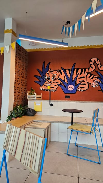 This is an interior photograph of a brightly lit, modern cafe or restaurant. The dominant feature on the back wall is a large, colorful mural of a tiger depicted in a cartoonish style, with bold orange and black stripes and large blue foliage. Below the mural, there is a ledge with white tiled backsplash, and on this ledge, there are some potted plants and wooden crates. Two small, round black tables with slender legs are visible. To the right, a blue-framed chair with a wooden seat and cushion is positioned at one of the tables. In the foreground, there are more wooden furniture elements, possibly seating or tables, with a blue frame structure in the lower left corner. The lighting appears to be a combination of overhead fixtures, including a long, blue, rectangular light. Decorative triangular flags in blue and white are strung across the ceiling. The overall impression is a vibrant and playful space. This image was taken in the city of Vladimir, Russia.