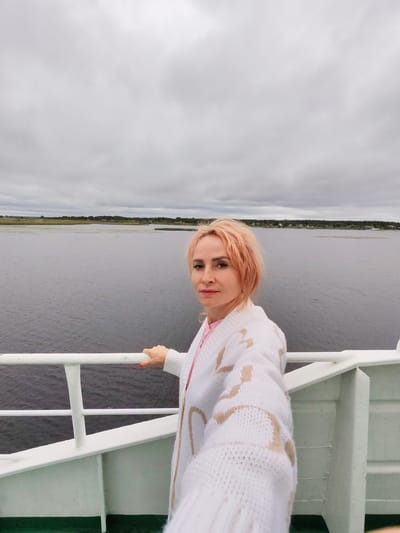 A woman with short, peachy-pink hair and fair skin is taking a selfie on a white ferry. She is wearing a white knit cardigan with a tan pattern over a pink top. Her left hand is resting on a white railing, and her fingernails are painted red. The background is a large body of dark water, with a distant, green, tree-lined shore dotted with small houses. The sky is overcast with gray clouds. This scene is likely on the Neva River or Gulf of Finland, near Shal'skii, Russia, given the landscape and ferry.