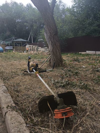 The image depicts a yard in Rassvet, Kazakhstan, with a string trimmer resting on dry grass and debris. The yard appears to have recently undergone landscaping or clearing, as evidenced by the cut vegetation scattered on the ground. In the background, a wooden gazebo with a green roof stands next to a dark brown fence. A ladder leans against a tree, and construction materials are visible near the gazebo. A dark-colored car is parked to the left of the gazebo. The overall scene suggests a residential property in a suburban or rural setting.