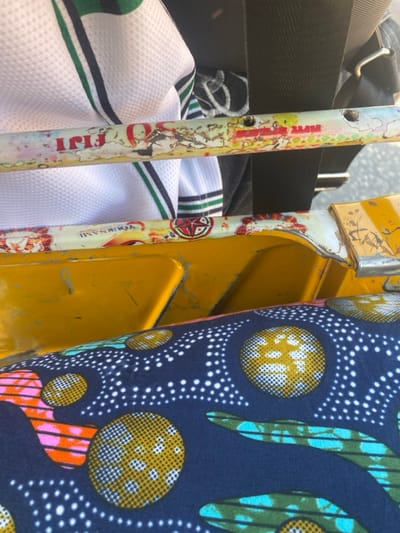 The scene presents a vibrant, cluttered interior. A yellow structure frames the view, beside it a pair of decorated poles, one bearing the faded "Fiji" label and another "YourName". A white shirt with green stripes adds to the disarray. A bold, patterned textile features circles with white dots on a dark background, juxtaposed against geometric patterns in green, orange, and pink. A black bag hangs overhead, completing the compact composition.