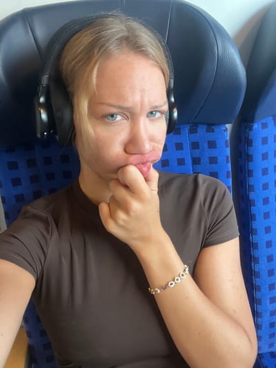 A person is lying on their side on a blue upholstered seat, likely on public transportation, as indicated by the patterned seat fabric and the dark blue headrest. The person is wearing a brown t-shirt and headphones, and has a bracelet with small flower-shaped charms on their right wrist. Their left hand is touching their face, with their index finger resting on their lips, and their tongue is visible. The person has light-colored hair and blue eyes. The lighting suggests it could be daytime. The image seems to be a selfie, with the camera positioned above and to the side of the person. There is no visible text in the image. The scene conveys a sense of casualness and perhaps boredom or playfulness. The location is identified as Ampfing, Germany.