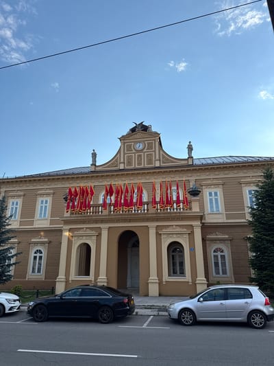 The image captures the National Museum of Montenegro, located in Cetinje, Montenegro. The building is an imposing, two-story structure with a beige facade and ornate architectural details, including arched windows and decorative cornices. A prominent balcony on the upper floor is adorned with numerous red flags, each featuring a golden emblem. Above the balcony, a clock face is visible on the building's central pediment, flanked by two statues. The sky above is a clear blue with a few scattered clouds. In front of the museum, several cars are parked along the street, including a black sedan and a silver hatchback. A tall evergreen tree stands to the right of the building, and another evergreen is partially visible on the left. The scene suggests a daytime setting, possibly a special occasion given the display of flags.
