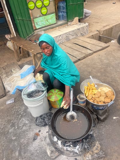 The image depicts an outdoor street food stall in Chikaji, Nigeria, focusing on a person engaged in deep-frying. The central figure is an individual, likely female, wearing a teal headscarf (hijab) and a matching top or abaya. She is shown in a squatting or low-seated position, holding white, peeled pieces of food, possibly plantain or yam, and is in the process of adding them to a large, dark frying pan filled with hot oil. A metal slotted spoon or skimmer is also visible in the pan.
The cooking station comprises the large frying pan situated over an open charcoal fire, with glowing coals and ash visible beneath. To the left of the pan, a sizable metal bowl contains a fresh batch of golden-brown fried plantain slices and cubed, light brown fried tofu or soybean curd (wara/awara). Below this, a smaller metal strainer bowl is present, along with a small transparent container with a white lid, possibly for ingredients.
Near the cook, a large white plastic bucket holds more unfried food pieces and some black plastic bags. A green and brown striped plastic basin is also on the ground. The immediate surroundings consist of an uneven, rough ground surface, possibly concrete or dirt, characteristic of an informal market setting.
In the background, to the left, green metal shelving units display numerous plastic bottles of water, clearly branded "oramo." Several green circular mats or coasters, also bearing the "oramo" logo and the phrase "KEEP EXPLORING," are visible. Partially visible in the distance are the legs of another person in light trousers. The scene, illuminated by natural light, portrays a typical daytime street food preparation and vending activity in the region.