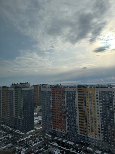 The image depicts a densely populated urban landscape in Анкудиновка, Russia. High-rise apartment buildings dominate the right side of the frame, characterized by repetitive windows and varying color schemes (grey, yellow, orange, green). A network of roads and parked cars, partially covered in snow, are visible at the base of the buildings. The left side of the image shows a cloudy sky with hints of sunlight breaking through. The image is vertically oriented, presenting a high-angle view of the scene.