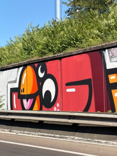 This image depicts a section of a bridge or overpass with graffiti art. The graffiti features a cartoonish character with large white eyes outlined in black, and an orange beak-like mouth with pink accents. The character appears to be on a red background, with parts of orange and white shapes visible above it, possibly part of another design. The graffiti is applied to what appears to be metal panels.
To the left of the graffiti, there is dense green foliage, suggesting vegetation growing along the side of the structure. A portion of a light-colored pole or support structure is visible in the upper left corner, against a clear blue sky. To the right of the graffiti, there is a concrete barrier and a paved surface, likely a road or walkway. The overall scene suggests an outdoor environment, possibly an urban or suburban setting where infrastructure meets nature. The lighting indicates it is daytime, with sunlight casting shadows.
There are some visible text elements in what appears to be Japanese characters in pink on a white background at the top of the graffiti, to the left of the red section. These characters are partially obscured. The graffiti itself has bold black lines and bright colors, giving it a vibrant and somewhat playful appearance. The image is captured in portrait orientation.
