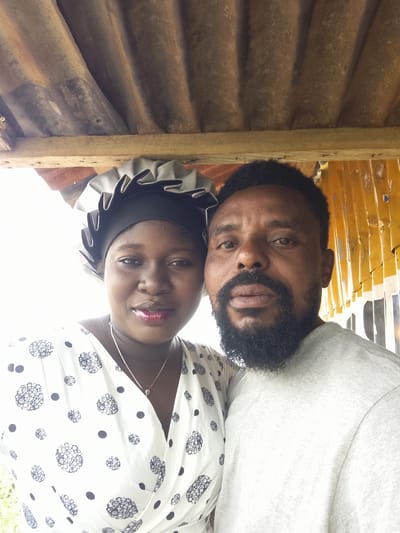 This media file is a selfie featuring a man and a woman. They are positioned close together, with the man on the right and the woman on the left. The man has dark skin, a beard, and is wearing a light gray t-shirt. The woman has dark skin and is wearing a white dress with a black circular pattern and a black headscarf. Both subjects are looking at the camera.

They appear to be outdoors, under a rustic shelter with a corrugated metal roof supported by wooden beams. The background to the right shows a yellow corrugated metal structure, possibly part of a building or fence. The lighting suggests it is daytime, possibly overcast given the diffused light.

The scene is informal, with the couple taking a selfie together. Their expressions are neutral, but the close proximity suggests a personal relationship. There is no visible text in the image. The setting appears to be a rural or less developed area, consistent with the provided location context of Agwala, Nigeria. The overall impression is of a candid moment captured between two individuals in an outdoor, sheltered environment.
