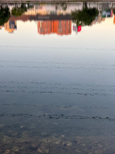 The image captures a reflection on water, likely a river or canal, in Reutov, Russia. The scene is divided horizontally. The left side of the image shows the murky, shallow bottom of the body of water, with stones and debris visible beneath the surface. The water is dark and somewhat opaque.
The right side of the image displays a distorted reflection of the surrounding environment. Buildings, possibly apartments or office blocks, are reflected as elongated, textured shapes. One building has a prominent orange or red facade, while another appears to have a red rooftop or sign. Trees and greenery are also reflected, appearing as blurred masses of dark green. A pathway or embankment with some railing or fencing is visible along the edge of the reflected scene. The reflection is wavy and fragmented due to the movement of the water, creating an abstract and painterly effect.
The sky in the reflection is a pale, hazy white or light grey, suggesting it is either overcast or possibly late evening or early morning. There is no clear indication of a specific time of day, but the soft light might suggest twilight. The overall mood is serene and contemplative, with the natural texture of the water interacting with the man-made structures. No people are visible in the image, and no text is discernible.