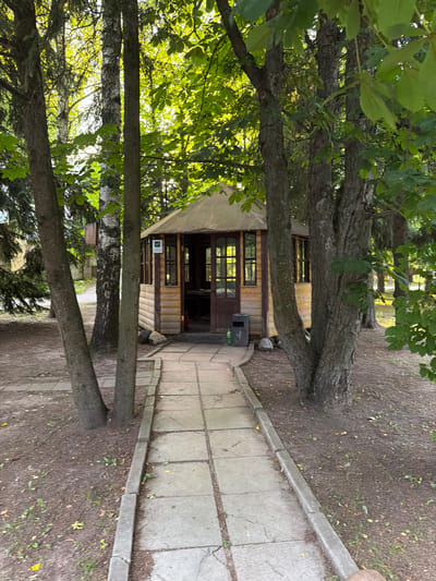 The image depicts a path leading to a small wooden building with windows. Trees with green foliage are prominent in the foreground and frame the scene. The path is made of concrete slabs and is bordered by a concrete curb. The ground around the path is covered with dirt, leaves, and some sparse grass. The building appears to be a simple structure, possibly a shed or a small kiosk, with wooden siding and a dark-colored door. There are windows on the front and side of the building. A trash can is visible next to the entrance. In the distance, more trees and greenery suggest a natural or park-like setting. The lighting indicates it is daytime, with sunlight filtering through the leaves. The overall impression is one of a peaceful outdoor location. The orientation of the image is rotated 90 degrees counterclockwise from standard landscape.