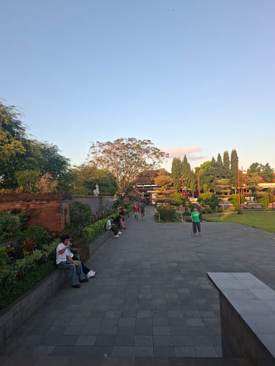 The image depicts an outdoor scene in the city of Gianyar, Indonesia, likely during late afternoon or early evening based on the lighting. The foreground features a wide paved pathway with a textured, tiled surface. To the left, a low, brick wall is covered with lush green plants and colorful foliage, suggesting a garden or manicured landscape. Several individuals are seated on the edge of this wall, some interacting or resting. One man in a white t-shirt and jeans is holding a microphone and wearing headphones, possibly performing or engaged in a recording.
Further back, the pathway leads towards a large, traditional-looking building with dark wood and tiered roofing, suggesting it could be a temple or cultural site, characteristic of Balinese architecture. The area around the building is a grassy park with numerous trees, including tall, slender ones and broader, leafy trees. Several people are visible walking or standing in this area. One man in a green shirt is standing with a cane, looking towards the building. The sky is clear and light blue with a hint of pinkish clouds near the horizon. A single bird is visible in flight against the vast expanse of the sky. The overall impression is a public space, possibly a tourist attraction or a gathering place, with a blend of architectural elements and natural greenery.