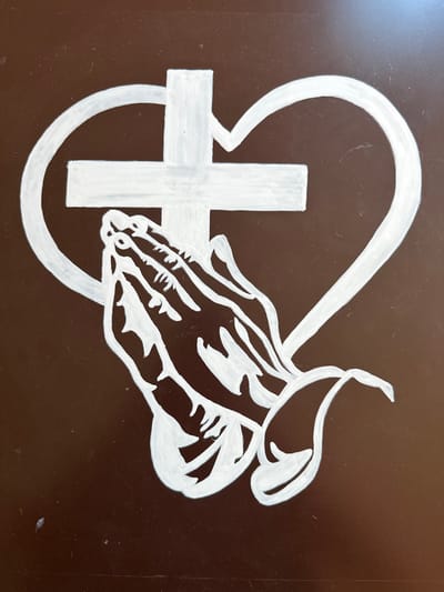 This media file depicts a white drawing on a dark brown background. The drawing features a Christian cross inside a partially formed circle, with a stylized representation of praying hands superimposed over it. The hands are depicted in profile, with fingers intertwined, suggesting reverence or supplication. A curved line arcs over the cross and hands, possibly representing a halo or divine protection. The overall composition evokes themes of faith, prayer, and spirituality. There is no visible text in the image. The setting is likely an indoor surface, such as a wall or plaque, given the flat, dark background and the scratch marks visible on its surface. The style of the drawing is somewhat simplistic but expressive, with bold white lines defining the shapes. The specific location context of Svilengrad, Bulgaria, does not offer any visual cues within the image itself, but suggests a possible cultural or religious significance of the artwork in that region.