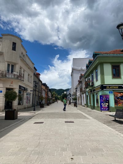 The image shows a street scene in the city of Cetinje, Montenegro. The street is paved with light-colored stone tiles and lined with buildings on both sides. The buildings are of varying architectural styles, with some having a more traditional appearance and others appearing more modern. Many of the buildings house shops and businesses, indicated by storefronts and signage, such as "step sport" and "ZLATNIK". There are several streetlights and planters with small trees along the sidewalks. A few people are visible walking on the sidewalk. In the distance, the street curves and leads towards a mountainous landscape under a partly cloudy sky. On the right side of the street, a green building with a tiled roof features a sign that reads "ZLATNIK" and a poster advertising "ZLATNI KLUB". A dark-colored park bench is also present on the right sidewalk.