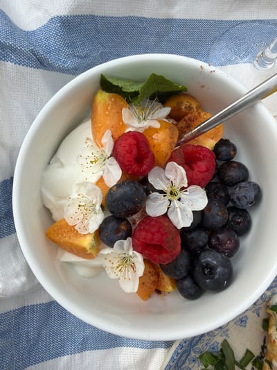 A white bowl is filled with a yogurt base, topped with raspberries, blueberries, and segments of yellow-orange fruit, garnished with white flowers. A silver spoon is partially submerged in the contents of the bowl. The bowl is set on a white and blue striped cloth. Part of a plate with food is visible in the upper right corner. The location is Gangloff, Germany.