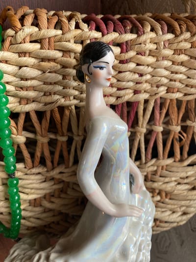 A porcelain doll, depicted in a sideways orientation, rests on a woven basket. The doll has dark hair styled in a bun, blue eyes, red lips, and is wearing a pearlescent white gown that reflects iridescent colors. A gold earring is visible on her left ear. Below the doll, a strand of green beads is visible, possibly jade. The basket itself is intricately woven from natural fibers, with variations in color from light beige to a reddish-brown. The texture of the weaving is rough and organic. The scene appears to be indoors, possibly in a home or a display setting, given the lighting and focus on the objects. There are no discernible clues about the time of day, weather, or specific location beyond the provided context of Zelenodol'sk, Russia. The doll's pose and serene expression do not convey a specific emotion or activity, suggesting it is a static arrangement rather than a depiction of an event. No text is visible in the image.