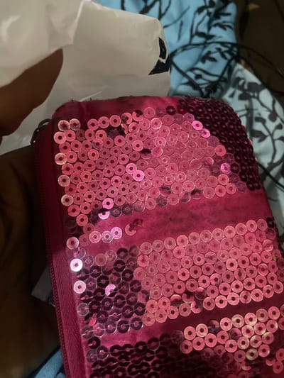 The image displays a close-up of a pink sequined wallet, partially obscured by a white paper bag. The wallet has a zipper closure and is covered in small, circular pink sequins. In the background, a blue fabric with a black floral pattern is visible. The lighting suggests an indoor setting. There is no discernible information about the location in Malete, Nigeria, as the image focuses solely on the object and its immediate surroundings.