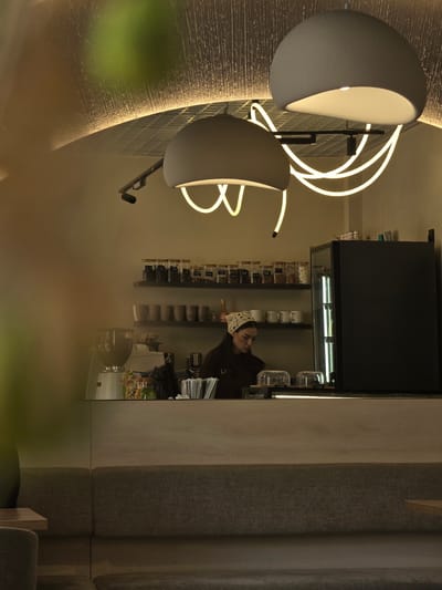 The image shows the interior of a cafe in Pyatigorsk, Russia. A woman with a patterned headscarf is working behind a counter. In front of her are two glass domes, a coffee grinder, and various bottles and jars on shelves. A black refrigerator is to her right. Above the counter, two large pendant lamps hang from the ceiling, and a neon light fixture forms a curved pattern. The cafe has a modern design with a textured ceiling and built-in seating. The overall lighting is warm and ambient.