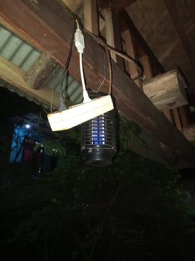 The scene depicts a nighttime outdoor or semi-outdoor environment in Naya Andura, India. A black, cylindrical insect zapper, emitting a blue internal light, is centrally positioned and suspended below a wooden beam. Power to the zapper is supplied via a black electrical cord plugged into a white, multi-outlet power strip, which is also suspended from or secured near the wooden beam. A white power adapter is plugged into an adjacent outlet on the same power strip, and a long, light-brown electrical cable is draped over the wooden beam. Above and to the left, corrugated metal roofing is visible. Additional wooden structural elements, including a small wooden box-like structure, are observable in the upper right quadrant. Below and in the background, dense, dark green foliage is present. A faint blue light emanates from an unidentified structure in the distant background. No human subjects are depicted.