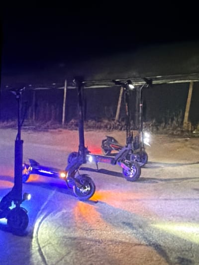 In Ognianovo, Bulgaria, at night, four electric scooters are parked under a covered structure. The scooters are equipped with bright LED lights that illuminate the asphalt surface with vibrant blue and orange hues. The area appears to be an outdoor space, possibly a parking lot or an area with a canopy, with dimly lit vegetation visible in the background. The lighting suggests a deliberate setup, perhaps for aesthetic display or to draw attention to the vehicles.