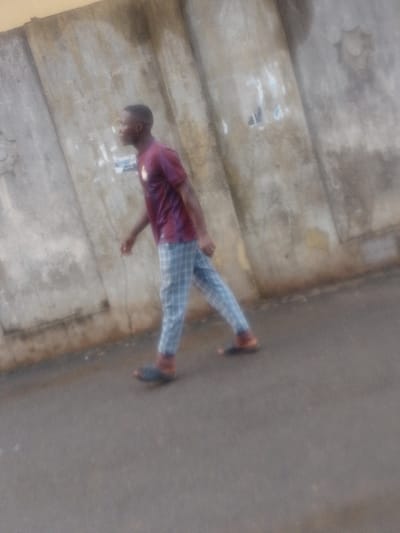 A person wearing a maroon t-shirt, blue plaid pants, and dark sandals is walking on a paved surface. The individual is moving from left to right in the frame, with a concrete wall visible in the background. The wall is light gray and appears slightly weathered. The image has a somewhat hazy or soft focus quality.