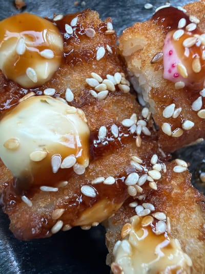A close-up, overhead shot focuses on a dish of food, possibly sushi or a similar appetizer, arranged in a black container. The food consists of several pieces of crispy, golden-brown fried items topped with creamy white sauces and a dark, glossy sauce, possibly teriyaki or a similar glaze. White sesame seeds are liberally sprinkled over the entire dish, adding texture and visual appeal. Some pieces of the food are slightly broken, revealing layers of rice and filling. One piece has a small dollop of bright pink sauce with red specks on it. The background is dark and out of focus, suggesting the food is the sole subject of interest. There are no people or any discernible features of the city of Dyurtyuli, Russia, in the image.