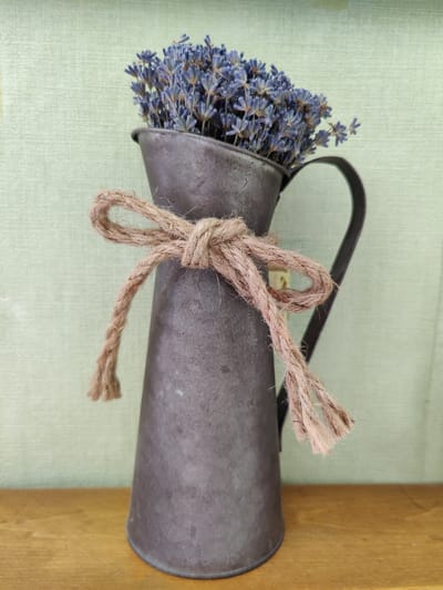 A close-up, vertical shot shows a rustic bouquet of dried lavender flowers in a dark, metallic jug. The jug has a handle and is tied with a rough, brown twine bow around its neck. The jug and its contents are placed on a light brown wooden surface. In the background, a light green, textured surface creates a simple backdrop. The overall aesthetic suggests a peaceful and natural setting, possibly a home or a craft display. The lighting appears to be natural and diffused, creating a soft ambiance. There is no visible text in the image.