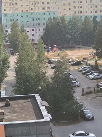 An aerial view of a residential area in Nizhnevartovsk, Russia, shows a multi-story apartment building with alternating bands of yellow, mint green, and white. In front of the building, a paved area functions as a parking lot with numerous cars parked. A playground with slides and swings is visible, adjacent to a sand-filled volleyball court with a net. Mature trees are scattered throughout the scene, casting shadows on the ground. In the foreground, the flat roof of a lower building with a chimney is visible. The sky is overcast, indicating a daytime scene.