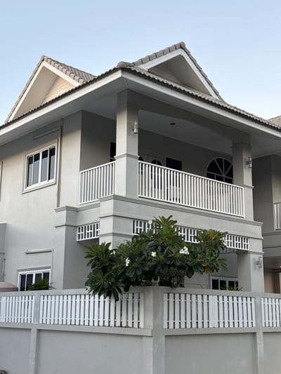 A light gray two-story house with a gray tiled roof is shown from a low-angle outdoor perspective. The house has a white railing balcony on the second floor. A white wooden fence runs along the front of the property, with a smaller white fence above a gray concrete wall. Green trees with white flowers are in front of the house. The sky is clear and light blue. This image was taken in Baan Harn, Thailand.