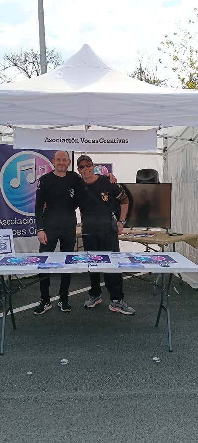 An outdoor scene in Ripollet, Spain, shows two adult males standing behind white folding tables under a white canopy tent. The tent's back wall features a banner reading "Asociación Voces Creativas" and a large circular logo with musical notes. The male on the left, with short hair and a slight beard, wears a black long-sleeve shirt with a white logo on the left chest, dark trousers, and black-and-white athletic shoes. The male on the right wears a black baseball cap, dark sunglasses, a black short-sleeve shirt over a striped long-sleeve shirt, dark trousers, and grey-and-orange athletic shoes; he also wears a black crossbody bag. His left arm is around the shoulder of the male on the left. Both individuals are smiling. The white folding tables in the foreground display multiple brochures and a stand with a QR code, all featuring the "Asociación Voces Creativas" logo. Behind these tables, a larger table covered with a brown cloth holds a black flat-screen monitor and a black speaker. The ground beneath the tables is dark asphalt. Above the tent, bare tree branches and a grey utility pole are visible against a partly cloudy sky.
