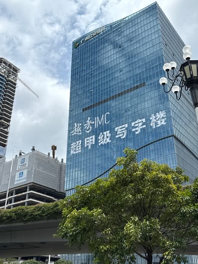A high-angle, medium shot captures a modern cityscape in Guangzhou, China, focusing on the Huangpu District. The primary subject is a tall, glass-facaded skyscraper with white lettering across its surface. The lettering appears to be a slogan or advertisement. To the left of this skyscraper, another building under construction is visible, with its structural framework exposed and a crane extending upwards. In the foreground, lush green trees obscure the lower portions of the buildings and an elevated roadway. The sky is overcast with clouds, suggesting a temperate climate. The scene is devoid of any human presence.