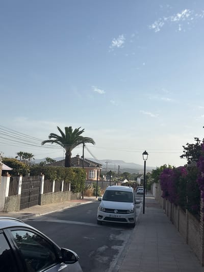 In Cañada De La Plata, Spain, a street scene unfolds under a clear blue sky with sparse clouds. The street, paved with asphalt, slopes gently downhill and is lined with a sidewalk made of gray rectangular tiles. On the left side of the street, a black metal gate is set into a stone and white pillar fence, bordered by dense green bushes. Beyond the fence, a house with an orange roof and pale orange walls is visible, featuring a bay window. A prominent palm tree stands tall near this house, its fronds spread wide against the sky. Overhead, numerous power lines crisscross the scene.
Parked on the right side of the street is a white Volkswagen Caddy van, its license plate reading "1429 KJS". Further down the street, another white car is parked. On the right sidewalk, a traditional black lamppost stands sentinel. A brick wall covered in vibrant purple flowering plants runs along the right side of the sidewalk. In the foreground, the reflection of trees and the sky can be seen in the dark paint of a parked car. In the distance, rolling hills and mountains form the horizon, with a hint of a construction crane visible. The overall impression is a quiet residential street in a warm climate.