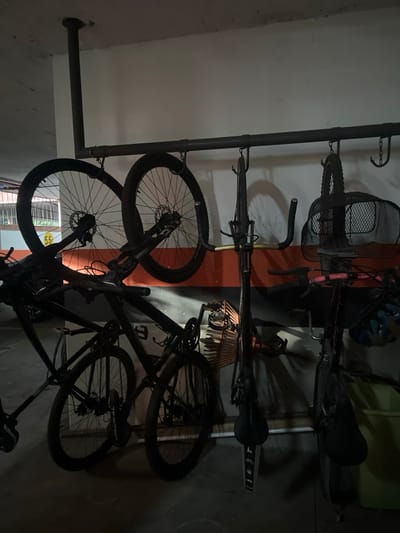 This image depicts a bicycle storage area within an underground parking garage or similar indoor facility located in Bogota, Capital District, Colombia. The scene is illuminated by artificial light, casting shadows on the off-white concrete walls, which feature a horizontal orange stripe at mid-height. A dark grey concrete floor is visible.

A robust metal pipe rack system, L-shaped, is suspended from the ceiling and extends horizontally along the wall, serving as a multi-bicycle hanger. Several bicycles are stored:
- Two dark-colored bicycle wheels, consistent with mountain bike wheels featuring disc brakes, are hanging by their rims from the horizontal pipe on the left. A yellow "55" sticker is affixed to the left wheel.
- Three full bicycles are visible. Two are suspended vertically from the horizontal rack.
    - The middle suspended bicycle is dark, possibly black, with gold-colored tape on its handlebars and a "LIFE" sticker on its seat tube.
    - The rightmost suspended bicycle is also dark and features a black wire mesh basket attached to its front. A dark blue and white bicycle helmet is hanging near its handlebars.
- At least two additional dark-colored bicycles are parked upright on the floor beneath the hanging bikes. One, partially visible, has "JACKAL" printed on its frame and a light-colored, possibly wicker, basket attached to its front.

No people are present. The scene is static, showing an organized storage arrangement for multiple bicycles.