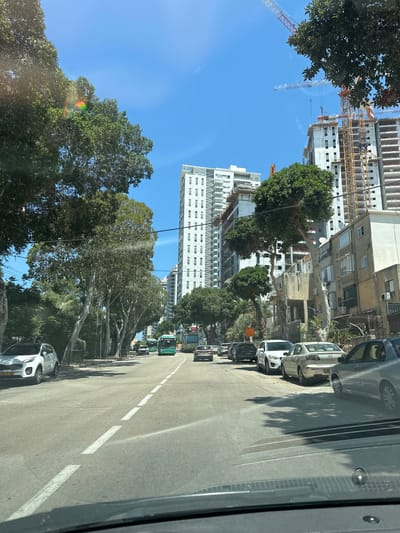 The image depicts a street scene in Haifa, Israel, on a clear, sunny day. The perspective is from inside a vehicle, looking forward along the road. On the left side of the street, a row of mature trees with lush green foliage lines the sidewalk. Behind the trees, a tall, modern white building with many windows dominates the skyline. Further in the background, a construction site is visible with several cranes and partially built high-rise buildings, indicating ongoing development.
The right side of the street is lined with parked cars, including a silver sedan, a white hatchback, and several other vehicles. The road itself is asphalt, with a white dashed line marking the lanes. Sunlight casts shadows of the trees onto the road. In the distance, more buildings and trees are visible, suggesting a densely populated urban environment. The time of day appears to be midday, given the bright sunlight and the length of the shadows. There is no discernible activity of people in the image, and no significant events are taking place. The overall impression is one of a typical day in a city with a mix of older and newer architecture, and ongoing construction projects.