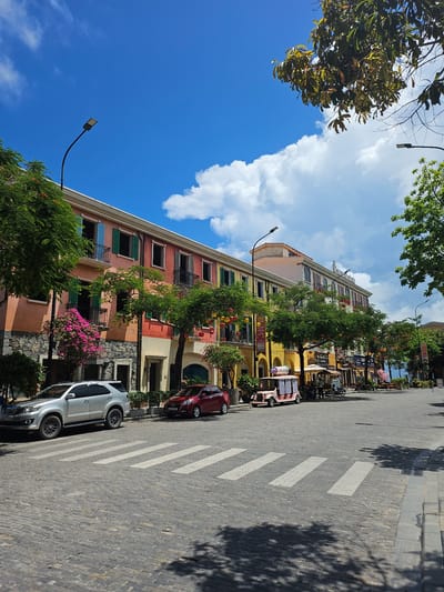 This is a landscape orientation, eye-level outdoor photograph taken on a sunny day, likely in the late morning or early afternoon, given the clear blue sky and bright daylight. The scene depicts a picturesque street lined with colorful buildings and lush greenery, suggesting a vibrant urban or resort setting.

The buildings are the dominant architectural features, displaying a variety of warm colors such as yellow, pink, and orange. They have multiple stories with arched windows and balconies, and some are adorned with shutters and decorative elements like hanging lanterns. Trees and flowering bushes with bright pink blooms are interspersed along the buildings and sidewalks, adding to the lively atmosphere.

In the foreground, a cobblestone street features a pedestrian crossing marked by white stripes. Two cars are parked on the street: a silver SUV closer to the foreground and a darker red sedan further down. Shadows of trees are cast across the street and sidewalk, indicating the position of the sun.

The sky is a vivid blue with scattered fluffy white clouds. The image is framed by the branches of trees, one with broad green leaves in the upper left corner and another with greener foliage in the upper right. The overall impression is one of a charming and well-maintained street in a warm climate, possibly a tourist destination. There is no discernible text in the image.