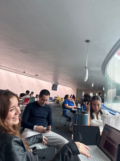 The image shows an indoor setting with several people seated at desks. The people appear to be students engaged in study or work activities. One person has a laptop open, and another appears to be looking at their phone. The location is likely a study area or library within a building. Based on the location details provided (Heroica Puebla de Zaragoza, Mexico), it's possible that this scene is taking place at a local university library or a study area in a public space. The lighting seems bright, possibly indicating natural light from windows. There are people seated further away, possibly other students or staff, and the general atmosphere appears to be one of quiet activity.
