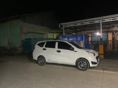 A white four-door hatchback is parked on the side of a paved road at night in Sungai Meriam, Indonesia. The car is facing to the right and appears to be in good condition. The background shows several buildings, including a corrugated metal structure and a building with a light blue and green facade. There is also a covered walkway with a metal roof and supporting pillars. A blue corrugated metal fence is visible between the car and the buildings. The road is dimly lit by artificial light, and the sky is dark, indicating it is nighttime. There are no people or animals visible in the image. The scene appears to be a street or an area outside of a commercial or residential property.