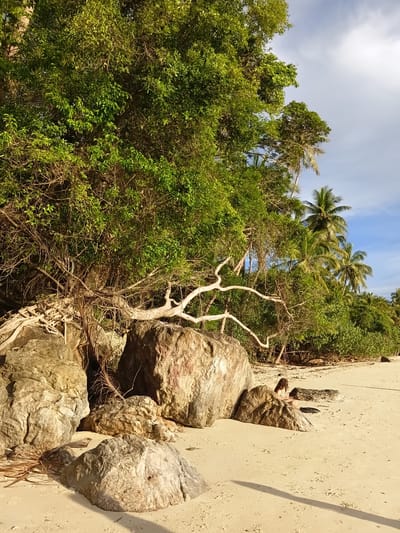 This image depicts a sandy beach adjacent to lush, tropical vegetation in Baan Pang Ka, Thailand. The foreground is dominated by several large, weathered rocks of various sizes, casting long shadows on the light-colored sand. A person is seated between two rocks in the middle ground, facing towards the right, with their back mostly to the camera. They appear to be a young adult with long, dark, curly hair, wearing a yellow and white patterned swimsuit. Behind the rocks and person, a dense wall of green foliage, including trees and palm trees, forms the backdrop. The sky above is a bright blue with scattered white clouds. The lighting suggests it is late afternoon or early evening, with the sun casting warm light on the scene.