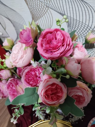 A close-up, eye-level shot captures a bouquet of artificial pink and rose-colored flowers. The flowers are densely arranged, with a variety of sizes and bloom stages, some fully open and others as buds. Green leaves and small green buds are interspersed among the flowers. The bouquet is presented against a textured wallpaper with a geometric pattern in shades of gray and beige. The lower portion of the image shows a dark red fabric, likely part of a vase or table setting, and a hint of a gold metallic edge. The lighting is even, with no distinct shadows. The scene appears to be indoors, on a flat surface. The location in Shakhtinsk, Kazakhstan, is not discernible from the image itself.