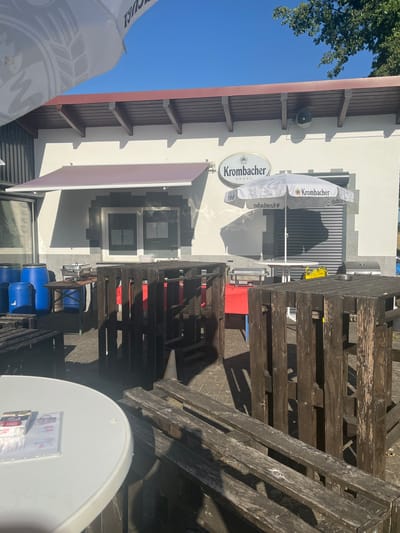 The image depicts an outdoor seating area in front of a building, likely a restaurant or cafe, in Niederwald, Germany. A white awning extends from the building above a window. To the left, several large blue barrels are stacked. In the center, a metal chafing dish is placed on a table, and to its right, a white umbrella with the "Krombacher" logo is open. Wooden pallet furniture, including tables and benches, is scattered throughout the foreground and midground. The ground is paved with light-colored bricks. The sky is a clear, bright blue, and sunlight casts shadows from the building and objects. A Krombacher sign is visible on the building. The scene suggests an establishment preparing to serve customers or currently operating in an outdoor capacity.