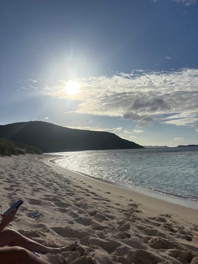 The image depicts a sunny beach scene, likely in the city of Olde Yard Village, British Virgin Islands. In the foreground, the lower legs of a person are visible, partially buried in light-colored sand. This person's left hand holds a dark-colored smartphone. To their right, a silver-colored beverage can is partially buried in the sand. The midground features a wide, curving sandy beach with numerous footprints, leading to a body of clear, light blue ocean water with sunlit ripples and gentle waves breaking at the shore. In the background, a large, dark green, tree-covered hill or mountain slopes down to the coastline, with a small, dark structure visible on its distant peak. Further right on the horizon, faint outlines of smaller landmasses are discernible. The sky above is bright blue with scattered white and grey clouds, and the sun is prominent and low, creating significant glare and lens flare.