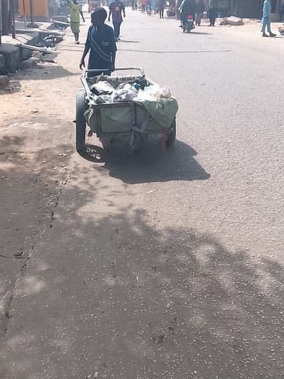 The scene depicts a sunlit urban street in Gawuna, Nigeria. In the foreground, a male individual, appearing to be an adult, with dark skin and dressed in a short-sleeved blue shirt with white horizontal stripes and dark trousers, is walking from left to right. He is positioned behind and pushing a two-wheeled metal handcart. The handcart is filled with assorted refuse, partially covered by a light green fabric, and exhibits signs of wear, including a seemingly flat or deflated left wheel. The ground consists of a rough asphalt road surface to the right, and a dirt/gravel shoulder to the left, separated by a raised concrete curb or drainage ditch containing scattered debris. Long, dark shadows, indicative of bright overhead sunlight, are cast on the road from the cart and the person. In the mid-ground and background, multiple additional pedestrians are visible walking on the asphalt road, alongside one motorcycle with a rider and passenger. Faint architectural outlines suggest urban or semi-urban structures lining the street in the distance.