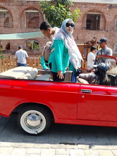 In Bejalen, Indonesia, a group of people are gathered around a bright red convertible. A man in a white t-shirt and jeans stands to the left, facing away from the camera. Next to him, a woman wearing a turquoise long-sleeved shirt and a patterned headscarf is looking at the camera, her face partially obscured by sunglasses. She is holding a brown bag. To the right, a person wearing a grey t-shirt and a black baseball cap is leaning into the convertible, speaking with another individual inside the car, who is wearing a grey t-shirt and a black baseball cap. The interior of the convertible features cream-colored seats. The background shows a rustic brick building with arched windows and a wooden fence. Sunlight illuminates the scene, casting shadows on the cobblestone ground.