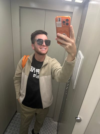 The image shows a man taking a selfie in what appears to be an elevator. He is wearing sunglasses, a black t-shirt with white text, and a light-colored fleece jacket. The elevator has metal flooring and light-colored walls. An orange object, possibly a bag or backpack, is visible on his shoulder. The man is smiling. There is text on the ceiling of the elevator, but it is too blurry to read. The overall lighting suggests it could be daytime. The setting, an elevator, is a common location for selfies. The man's expression indicates a positive emotion.
