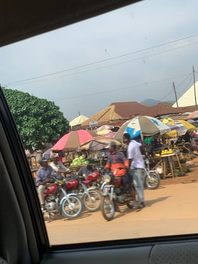The image shows an outdoor market scene in Dam Camp, Nigeria. Several motorcycles are parked in a line, some with people on or near them. Stalls with umbrellas are set up, displaying various goods, including fruits. A building with a sign that appears to say "PHARMACY" is visible in the background. The sky is overcast, suggesting it might be a cloudy day. The overall impression is a busy marketplace with activity and commerce taking place. The orientation of the image is rotated 90 degrees counter-clockwise.