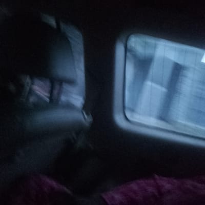 The image appears to be taken from inside a moving vehicle, possibly a car or bus. The location is Satellite Town, Nigeria, based on the available information. A blurred figure of a person is visible in the foreground, partially obscured by the headrest of the vehicle seat. They are wearing a pinkish-red garment. The focus is on the window to the right, showing blurred buildings and structures passing by, indicating movement. The lighting is dim, suggesting it might be evening or nighttime. The buildings visible through the window appear to be multi-story and box-shaped.