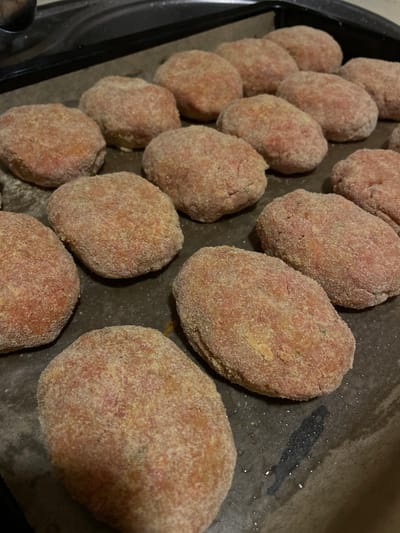 A close-up, overhead shot shows several uncooked, oval-shaped meat patties, each coated in breadcrumbs. They are arranged on a dark baking sheet lined with parchment paper. Some patties are in focus, revealing their pinkish interior and granular coating, while others towards the edges of the frame are softer in focus. The lighting suggests an indoor setting, possibly a kitchen, with a hint of shadow cast by the patties on the parchment paper. One shadow appears to be in the shape of a person. The scene is static, with no visible activity other than the preparation of food. There is no visible text in the image. The location context is La Línea de la Concepción, Spain, which provides a geographical setting for the domestic activity depicted.