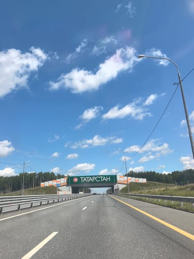 The image depicts a highway with a bright blue sky and scattered white clouds above. A pedestrian overpass spans the highway, featuring a green sign with white text that reads "ТАТАРСТАН" (Tatarstan). The overpass also has orange and white patterned sound barriers. A street lamp is visible to the right of the overpass. The highway is divided by a yellow line, and guardrails line the sides. Trees and vegetation are visible beyond the guardrails, suggesting a rural or semi-rural setting. The presence of the "Tatarstan" sign indicates the location is within the Republic of Tatarstan, Russia. The photo appears to have been taken from a vehicle moving along the highway.