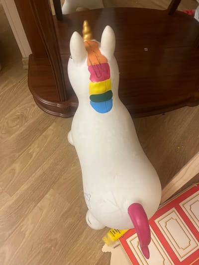 A white, plastic unicorn toy, painted with rainbow colors on its mane and a pink tail, sits on a wooden floor next to a dark wooden furniture piece. The unicorn is on its side, with its head facing away from the camera. There are some small pieces of yellow and red material on the floor near the unicorn, along with some small blue pieces scattered around. The image appears to be taken indoors in what looks like a domestic setting. No specific location within Edem, Russia can be determined from the image.