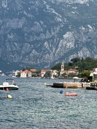 The image depicts a coastal scene in the town of Prčanj, Montenegro, characterized by a body of water in the foreground and a mountainous backdrop. In the foreground, the water is choppy, with several buoys of various colors (orange, yellow, and blue) scattered across the surface. A white motorboat with a black canopy is docked on the left side of the frame, bearing the registration "KT 1335". A small concrete pier extends into the water on the right, with a few people seated at tables and under an umbrella. The midground showcases a cluster of stone buildings with red-tiled roofs, indicative of traditional Mediterranean architecture. A prominent bell tower rises above the structures, suggesting the presence of a church. Green trees and vegetation surround the buildings. The background is dominated by a vast, rugged mountain range covered in sparse vegetation and rocky outcrops, creating a dramatic contrast with the tranquil waters and the quaint village. The overall impression is one of a serene coastal town nestled against a formidable natural landscape.