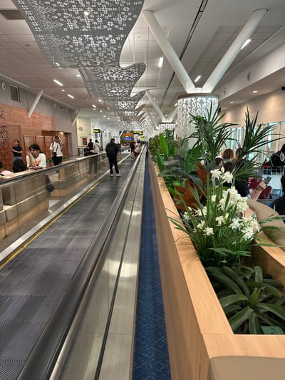 This is an interior shot of Kuala Lumpur International Airport (KLIA) in Sepang, Malaysia. A moving walkway extends into the distance, with a line of planters containing foliage and white flowers to the right. Passengers stand or walk on the walkway. To the left of the walkway, decorative panels hang from the ceiling. Beyond the walkway, lounge seating is visible. A signpost indicates directions. The lighting is bright and artificial.